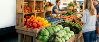 Customers shopping at farmers market produce stand with fresh vegetables and fruits displayed in wooden crates and boxes for sale.