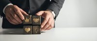 Male professional in dark business attire arranging wooden blocks with gold icons including heart, crown, and gear symbols, representing values, leadership, and business strategy on white surface.