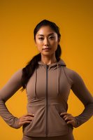 Young Asian woman in beige athletic hoodie posing confidently with hands on hips against vibrant orange background, expressing determination and strength.