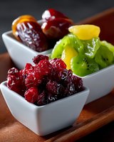 Colorful assortment of dried and candied fruits including cranberries, kiwi, dates and citrus pieces served in white ceramic bowls on wooden board.