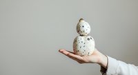 Caucasian hand holding stacked speckled eggs against neutral background, suitable for Easter, spring, and organic food concepts.