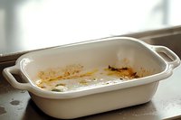 Empty white ceramic baking dish with food residue and grease stains after cooking, showing used condition on dark countertop in natural lighting.