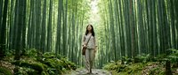 Woman with long dark hair, wearing casual beige and tan clothing, standing centered on a forest path through tall bamboo grove with lush green undergrowth.