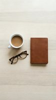 Flat lay of coffee cup, tortoiseshell glasses, and brown leather notebook on light wood surface, minimal workspace aesthetic.