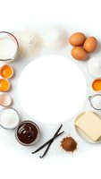 Fresh baking ingredients arranged around empty white plate on clean surface with flour, eggs, butter, milk, chocolate and vanilla beans for cooking.