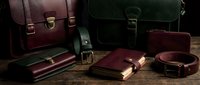 Luxury leather accessories collection featuring briefcases, wallets, belts and bags in rich burgundy and forest green colors on rustic wooden surface.