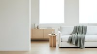 Minimalist living room with white sofa, sage green throw blanket, wooden dresser, and bright windows creating a calm, contemporary interior design space.