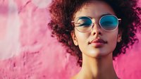 Young African American woman with natural curly afro hair wearing round sunglasses against pink wall background, expressing confidence and style in modern urban setting.