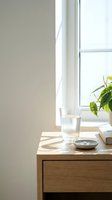 Minimalist wooden nightstand with glass of water and small bowl near bright window with green plant creating peaceful morning atmosphere in modern bedroom interior.
