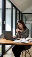 Young Asian businesswoman working on laptop at modern office desk with books and documents near large windows for remote work and productivity.