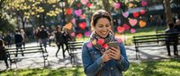 Young woman with curly hair using smartphone in autumn park with colorful foliage, smiling while texting outdoors during fall season.