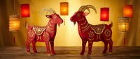 Red decorative goats with ornate patterns standing on wooden surface with hanging lanterns creating warm festive atmosphere for lunar new year celebrations.