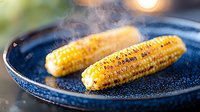 Grilled corn on blue ceramic plate with rising steam, golden kernels charred to perfection, butter melting on surface creates appetizing summer BBQ atmosphere.