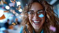 Young mixed race woman with curly hair and glasses laughing joyfully while colorful confetti falls around her against blurred bokeh background. Genuine happiness concept.