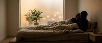 Woman with dark hair and medium skin tone lying on bed by window at sunset, gazing outside with contemplative expression, wearing casual clothing, with potted plant and warm bokeh lights visible in background.