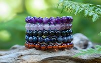 Colorful healing crystal bracelets stacked on natural wood with green bokeh background, featuring amethyst, rose quartz, and lapis lazuli beads for wellness and meditation.