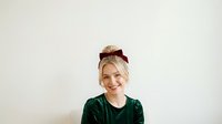 Young blonde woman with fair complexion wearing a dark green velvet shirt and burgundy bow headband, smiling at camera against neutral background for portrait and lifestyle use.