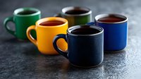 Colorful ceramic coffee mugs in green, yellow, navy blue and dark blue arranged in row on dark concrete background with steam rising from hot drinks.