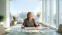 Confident mature businesswoman sitting at office desk with documents and charts, smiling at camera in modern corporate workspace with city view.