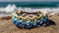 Handcrafted macrame bracelet with blue and beige crochet pattern on sandy beach with ocean waves in background.
