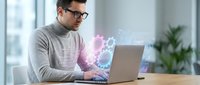 Young Caucasian businessman in glasses working on laptop with glowing digital brain hologram representing artificial intelligence and innovation in modern office.