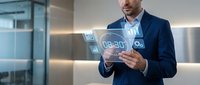 Male professional in dark business suit and light blue shirt holding tablet with glowing digital analytics icons and charts floating above screen in modern office environment.