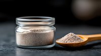 Ground brown powder in glass jar and wooden spoon on dark background. Natural organic seasoning or supplement for cooking and healthy nutrition.