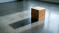 Wooden cube sitting on polished concrete floor in minimalist interior space with natural light casting sharp shadow.