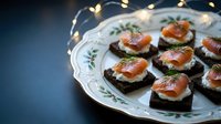 Elegant smoked salmon canapes with cream cheese and dill on dark bread served on vintage porcelain plate with festive lights creating warm holiday atmosphere.