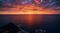 Dramatic ocean sunset with vibrant orange and red sky reflecting on calm water, rocky coastline in foreground, ideal for travel and nature backgrounds.