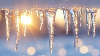 Glowing icicles hanging against blurred winter background with sunlight creating magical bokeh effect and warm golden light shining through crystalline ice formations.