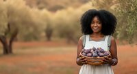 Young African woman with natural curly hair wearing white dress holding basket of fresh plums in olive grove orchard during harvest season.
