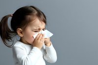 Young girl with pigtails blowing nose into tissue showing cold flu symptoms on gray background for healthcare medical concepts.