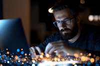Focused bearded man with glasses working late at night on laptop computer surrounded by warm glowing lights creating productive atmosphere for remote work.