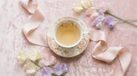 Elegant tea cup with herbal tea surrounded by soft pink ribbon and delicate flowers on textured pink background for feminine lifestyle and relaxation themes.