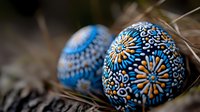 Decorative Easter eggs with intricate blue and gold floral mandala patterns painted on dark shells, nestled in natural dry grass. Traditional folk art design.