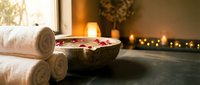 Spa wellness setup with rolled towels, flower petals in wooden bowl and warm candlelight creating relaxing atmosphere for massage and beauty treatments.