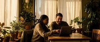 Young diverse couple working together on laptop at wooden table in cozy plant-filled home office with warm natural lighting during daytime.