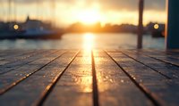 Wooden pier planks at sunset with golden sunlight reflection on wet surface, blurred marina and boats silhouettes in background creating peaceful harbor atmosphere.