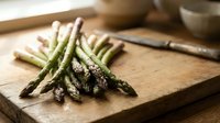 Fresh green asparagus spears on rustic wooden cutting board with knife, ready for cooking and meal preparation in modern kitchen setting.