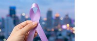 Female hand holding pink awareness ribbon against blurred city skyline at dusk, symbolizing health advocacy and support for charitable causes.