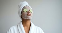 Young woman enjoying spa treatment with cucumber eye mask and facial cream wearing white bathrobe and towel for skincare and wellness routine.
