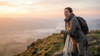 Middle-aged Caucasian male hiker wearing dark jacket and backpack, standing at wooden railing on mountain overlook at sunrise, gazing at misty valley landscape below during outdoor adventure and travel.