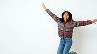 Young woman with long dark hair and East Asian ethnicity wearing colorful patterned jacket and jeans, jumping with arms spread wide in joyful celebration against white background.