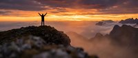Hiker celebrating success on mountain peak at dramatic sunrise with arms raised, overlooking misty valley and distant peaks in golden light.