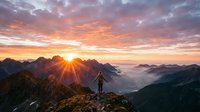 Hiker standing on mountain peak at sunrise with golden sun rays breaking through clouds and misty valleys below, adventure and nature exploration concept.