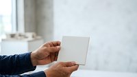 Man with medium skin tone holding blank white card or tile in hands, wearing dark blue sleeve, against blurred interior background for mockup and branding purposes.
