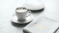 Steaming hot coffee in a white ceramic cup on marble surface with elegant gold lettering napkin, cozy cafe morning concept.