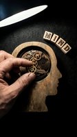 Adult male hand adjusting wooden gear mechanism inside carved head profile with mind text blocks, representing mental health, psychology, cognitive function and brain development concepts.