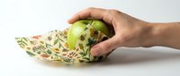 Hand wrapping a fresh green apple in reusable beeswax wrap with floral pattern, eco-friendly food storage concept on white background.